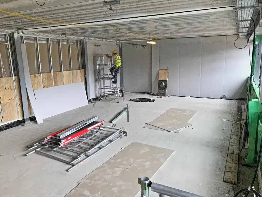 Construction worker on a ladder in an unfinished room with drywall, tools, and exposed ceiling beams.