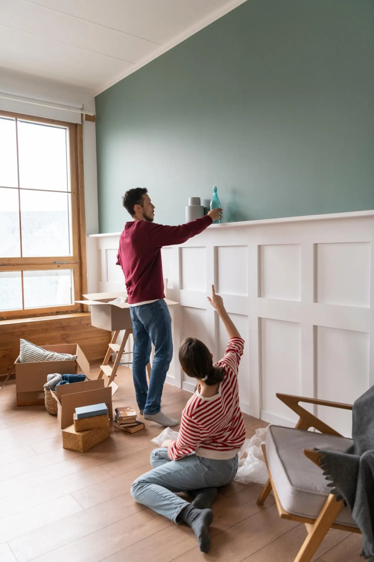 Couple decorates a room with boxes and furniture. Man reaches for decor on a shelf, woman points upwards.